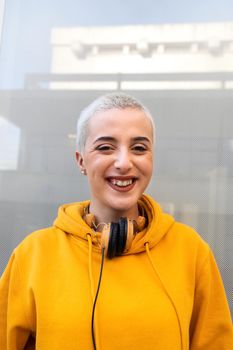 Vertical portrait of happy, confident teenage girl with short blond hair wearing orange hoodie and headphones looking at camera. Youth lifestyle and gen z concept.