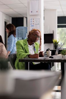 Employee in coworking open space, writing on clipboard, looking at laptop, sitting at workplace desk. Young african american woman working with company documents, taking notes