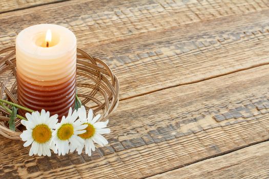 Burning candle and bouquet of chamomile flowers in wicker basket on wooden boards. Top view. Holiday concept.