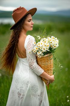 A middle-aged woman in a white dress and brown hat stands on a green field and holds a basket in her hands with a large bouquet of daisies. In the background there are mountains and a lake