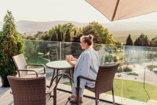 A middle-aged woman sits in a street cafe overlooking the mountains at sunset. She is dressed in a blue jacket and drinks coffee admiring the nature. Travel and vacation concept