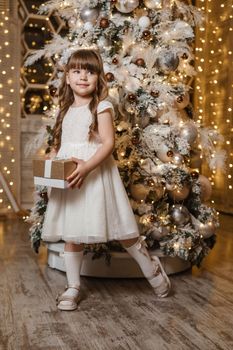 A girl in a festive light dress with a gift in her hands next to a Christmas tree, lights of garlands in the background. The concept of New Year holidays.