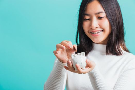 Asian beautiful young woman holding piggy bank on hands and putting coin, portrait relaxation of happy female smiling hold piggybank in palm, isolated on blue background, concept of saving money