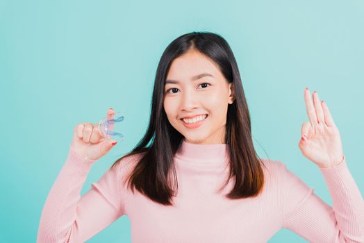 Portrait Asian beautiful young woman smiling holding silicone orthodontic retainers for teeth and show finger brackets saying OK sign, isolated on blue background, Dental hygiene healthy care concept