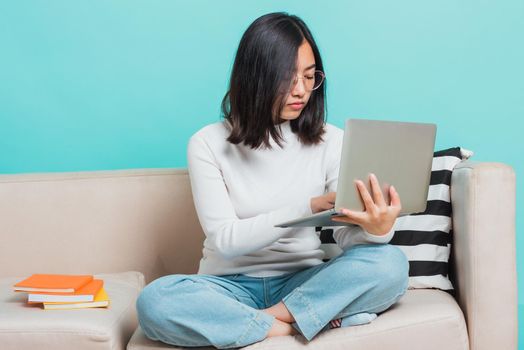 Asian beautiful young woman wear eyeglasses sitting on sofa using a laptop, portrait relaxation of happy female smiling in living freelancer working on computer studio shot isolated on blue background