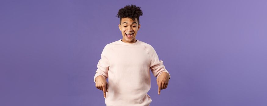 Joyful good-looking hipster guy with dreads rejoicing over good news, best deal ever in online store, favorite game finally in stock, pointing fingers down and rejoicing, purple background.