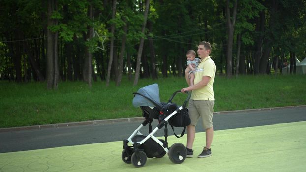 A young dad walks with his son in the park and carries a stroller in front of him. A man with a child in his arms is walking. 4k