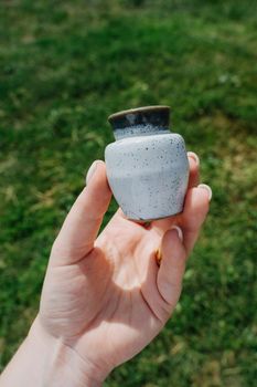 Small ceramic jug. The girl's hand holds a small clay jug of light gray color. Juicy green grass on the background. Vertical photo.