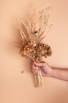 Bouquet of beige dried flowers, grass and leaves in woman hand on beige background top view