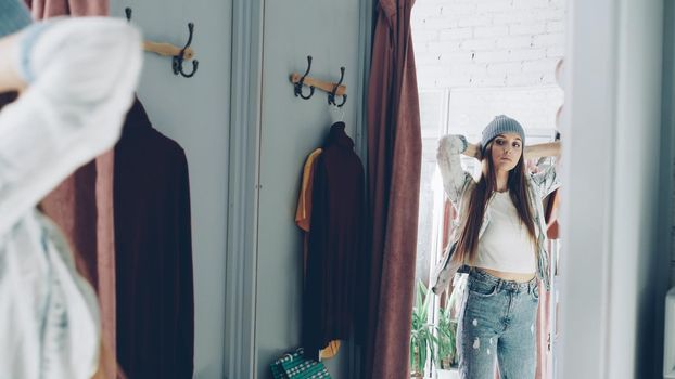 Mirror shot of young girl trying on clothes in fitting room. She is wearing denim jacket, jeans and hat, smoothing her hair, moving and looking in mirror to check image. Teenager shopping concept.