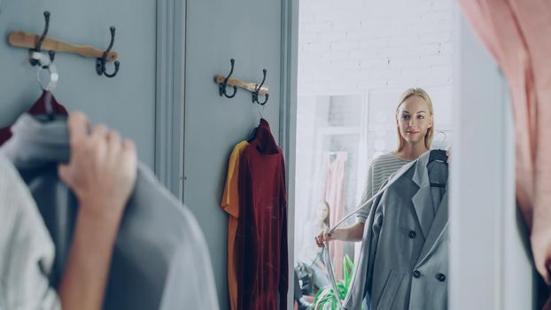 Attractive blond woman is trying elegant coat while standing in nice fitting room is clothing store. She is looking at herself in the mirror and smiling. Successful choice concept.