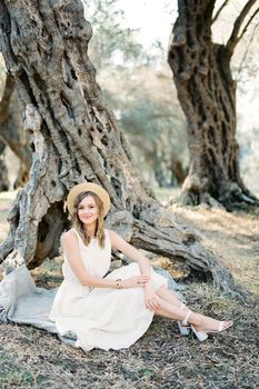 Young woman in a straw hat sits on a bedspread under an old olive tree. High quality photo
