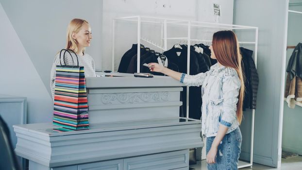 Cheerful customer is paying with credit card for purchased clothes while standing at cashier's desk.