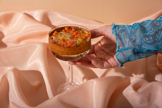 Still life from female hand and glass with water with mountain ash and dried leaf on folded fabric background. Minimalistic creative end of summer or autumn concept.