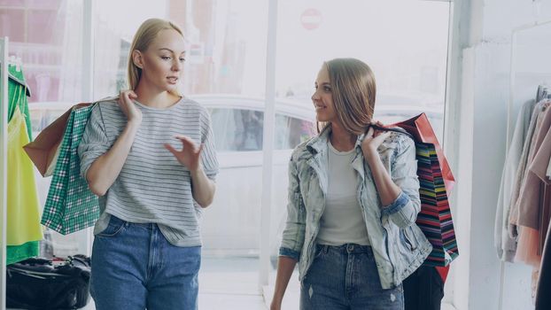 Two female friends are walking slowly through spacious boutique. They are holding numerous bright paper bags, chatting and looking at beautiful clothes and shoes on hangers and shelves.