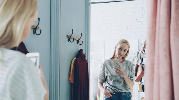 Pretty blond woman is making mirror selfie with smart phone while standing in nice fitting room in luxurious clothes boutique. She is posing, moving and smiling carelessly.