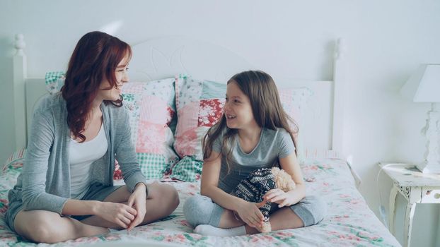 Young mother and little cheerful daughter talking and sharing secrets while sitting on bed at home in bedroom. Relationship and family concept