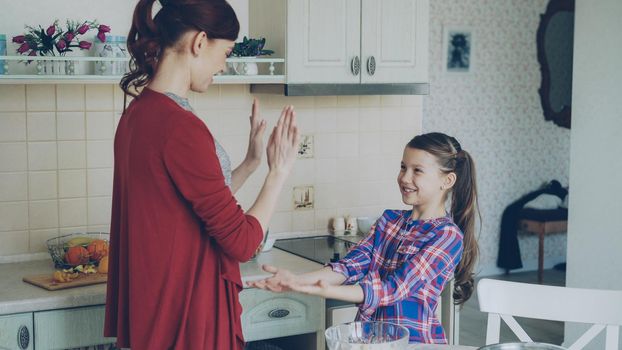 Happy young mother and cute funny daughter playing clapping hands game and have fun while cooking in kitchen at home. Family, cooking and people concept