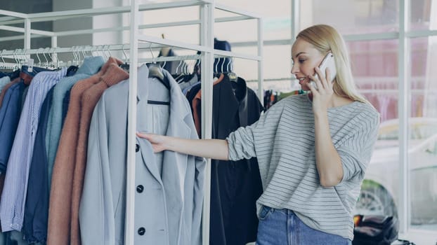 Cute blond girl is chatting on smart phone in modern boutique, touching clothes on hangers and smiling charmingly. Lots of fashionable clothes and large window in background.