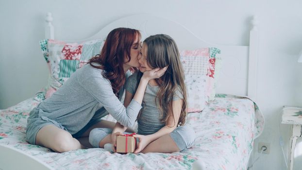 Young happy mother giving gift box to her little cute daughter embracing and kissing while celebrating child's birthday sitting on bed in light cozy bedroom at home.