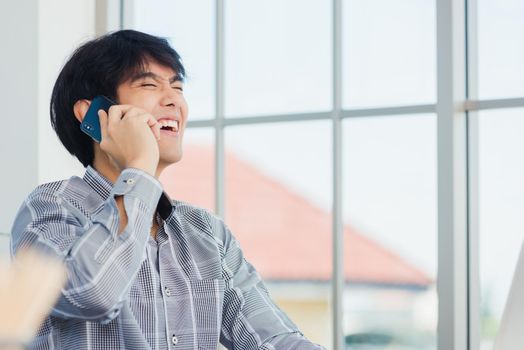 Asian businessman smiling and talking on a mobile phone at home office desk. The man smile call on smartphone with customer at the house, making sell offer