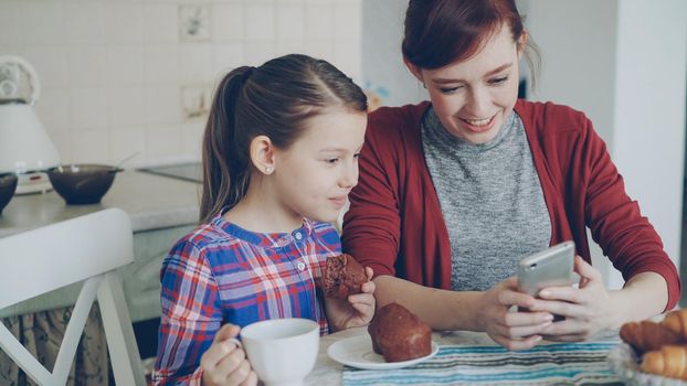 Cheerful mother and little daughter browsing smartphone together and smiling during breakfast in the morning in kitchen at home