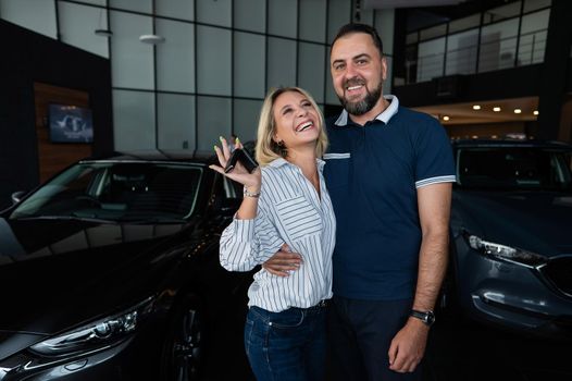 Happy caucasian couple hugging and woman holding new car keys in car showroom