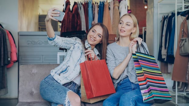 Attractive female friends are using smartphone to make selfie while sitting in women's clothing shop with lots of colourful paper bags. They are smiling, posing, laughing, gesturing.