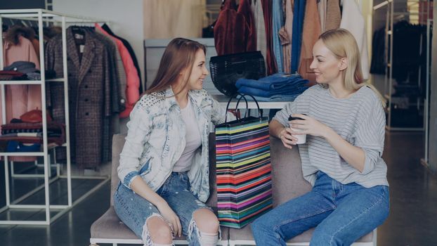 Two charming girls sitting in clothes shop with coffee, talking happily and laughing after shopping. Nice modern boutique with colourful garments, bags and shoes in background