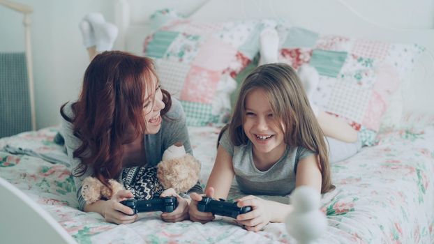 Happy young mother and cute daughter playing computer console games on TV while lying on bed at home in the morning and have fun