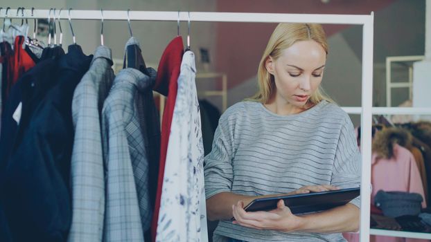 Young owner of clothing store is checking and counting garments on rails while working with tablet. She is inputting information about her goods. Successful small business concept.