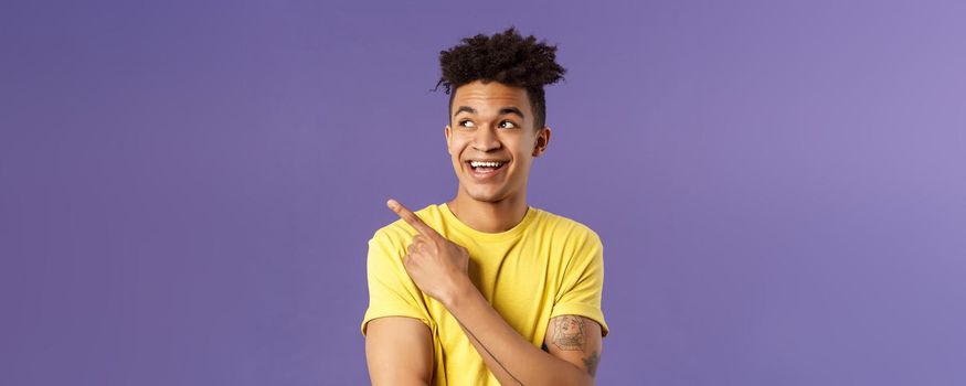 Close-up portrait of funny cute hispanic man with dreads, laughing and smiling at something, pointing looking upper left corner enthusiastic expression, purple background.