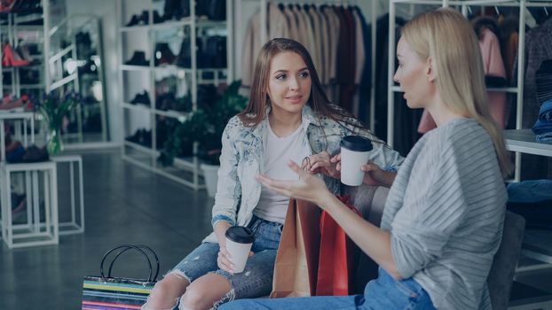 Two attractive girls sitting together and talking in spacious women's clothing store after long shopping. Stylish colourful garments, shoes and bags in background.