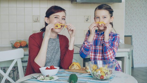 Joyful mother and cute cheerful daughter have fun grimacing silly with vegetables and looking into camera while cooking in the kitchen at home. Family, cook, and people concept