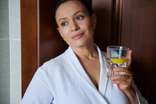 Close-up portrait of a charming middle aged woman in white bathrobe with a glass of refreshing lemon water for a good start to the day. Detox care for healthy skin and body. Anti-aging concept