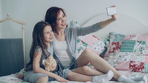 Happy mother and little girl taking selfie photo with smartphone camera and have fun grimacing while sitting in bright bedroom at home. Family, people and technology concept