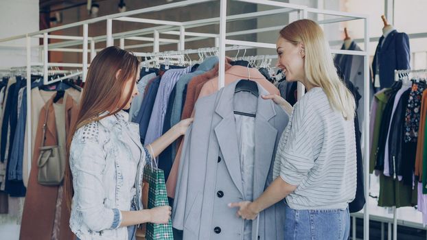 Young woman is helping her friend to find perfect coat. She is bringing her nice new model, telling about it, touching fabric and showing its advantages. Girls are talking and smiling.