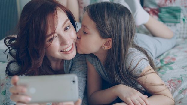 Closeup of Happy mother and little girl taking selfie photo with smartphone camera and kissing while lying in bright bedroom at home. Family, people and technology concept