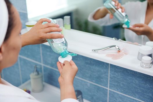 Selective focus on the hands of a blurred woman standing in front of a bathroom mirror and pouring a cleansing lotion on a cotton pad, to clean the face skin and remove make-up.