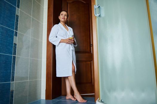 Full-length portrait of attractive middle aged woman in white bathrobe with a glass of refreshing lemon water for a good start to the day. Detox care for healthy skin and body. Anti-aging concept