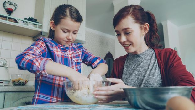 Little pretty girl helping her mother in the kitchen stirring dough for cookies into bowl. Mom and daughter have fun talking and laughing. Family, food, home and people concept