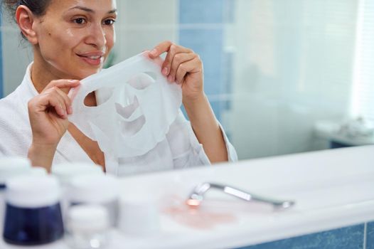 Selective focus on middle-aged multi-ethnic pretty woman, in white bathrobe, reflected in the bathroom mirror, holding a smoothing and moisturizing textile facial mask, taking care of her skin health