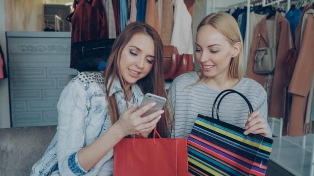 Cheerful young women chatting carelessly and using smartphone while sitting in nice clothing boutique. They are smiling and gesturing enthusiastically. Modern lifestyle concept.