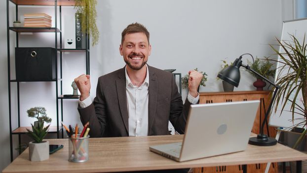 Concentrated businessman winner in suit working on laptop at home office desk typing browsing say Wow yes found out great big lottery win good news celebrate. Male manager freelancer man occupation