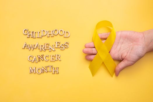 Childhood cancer awareness month text from wooden letters and female hand with yellow ribbon on yellow background.