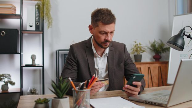 Businessman programmer software developer in suit working, making online purchase payment shopping with credit bank card and smartphone at modern office desk with laptop. Male freelancer business man