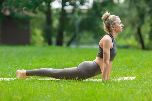 A slender woman in a gray top and leggings, standing on a sports mat in the summer, on the green grass in the park and doing yoga in snake pose. Copy space