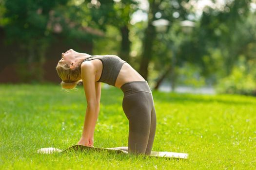 A slender woman in a gray top and leggings, standing on a sports mat in the summer, on the green grass in the park and doing yoga in the camel pose. Copy space