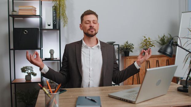 Businessman in suit working on laptop computer, meditating, doing yoga breathing exercise in lotus position at home office desk. Calm serene freelancer man taking break. Busy occupation. Peace of mind