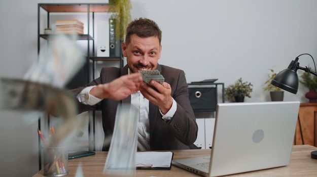 Cheerful rich businessman in suit working on laptop and wasting throwing money to camera, more tips earnings, big profit, success win lottery, shopping. Happy male freelancer man at home office desk